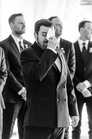 Emotional groom wiping tears at wedding altar in black suit, groomsmen standing behind in black and white photo.
