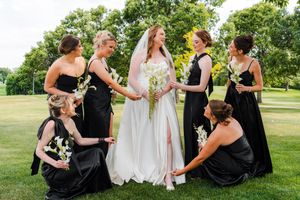 Bride in white gown with bridesmaids in black dresses holding white flowers on green lawn at outdoor wedding.