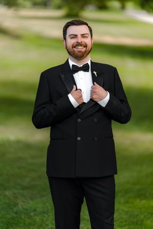 Smiling groom in black double-breasted tuxedo with bow tie and boutonniere poses outdoors on green lawn.