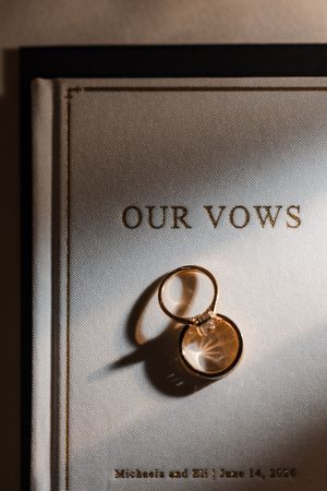 Gold wedding rings rest on a personalized vow book reading Our Vows for Michaela and Eli, June 14, 2026.