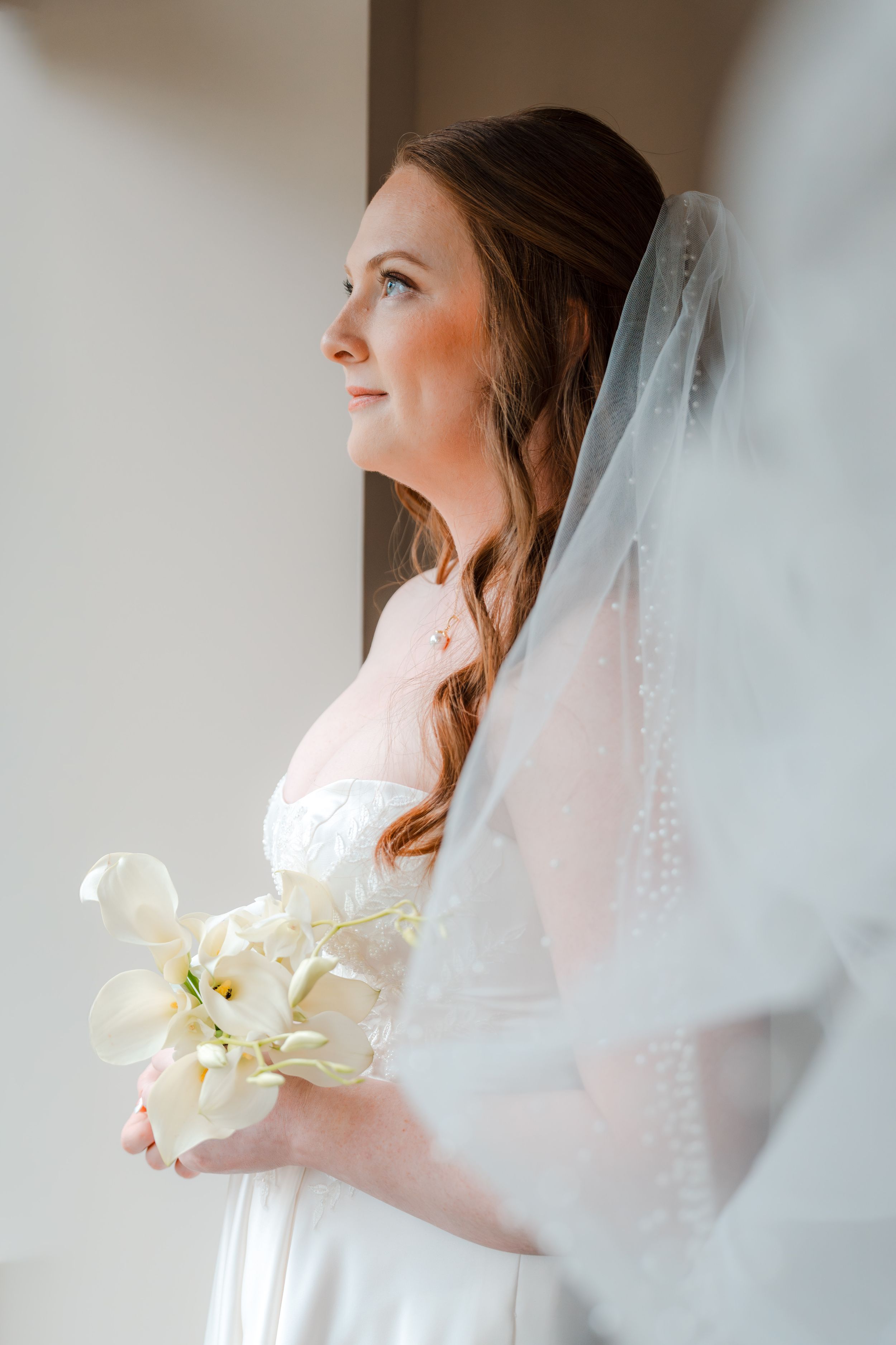 Bride in white gown and veil holds white calla lily bouquet, gazing softly toward natural light.