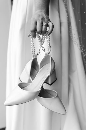 Black and white photo of a bride holding white block-heel shoes and pearl necklace in her ringed hand.