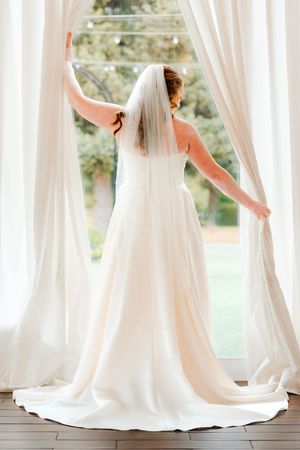 Bride in white gown and veil stands at large window with flowing curtains, viewed from behind.