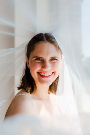 Smiling bride in white veil and dress poses near bright window on her wedding day.