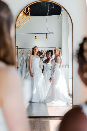 Three women in white wedding dresses posing together, reflected in an elegant arched mirror inside a bridal boutique.