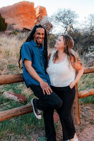 A laughing couple leans against a wooden fence with red rock formations in a scenic outdoor setting.