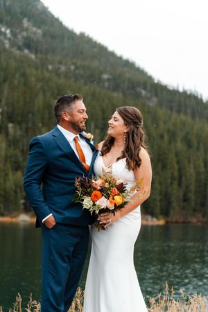 Bride and groom smile by a mountain lake, groom in navy suit with orange tie holding bouquet.