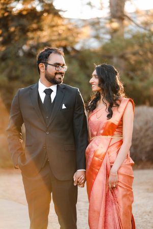Indian couple in traditional attire and black suit hold hands and smile outdoors in golden hour light.