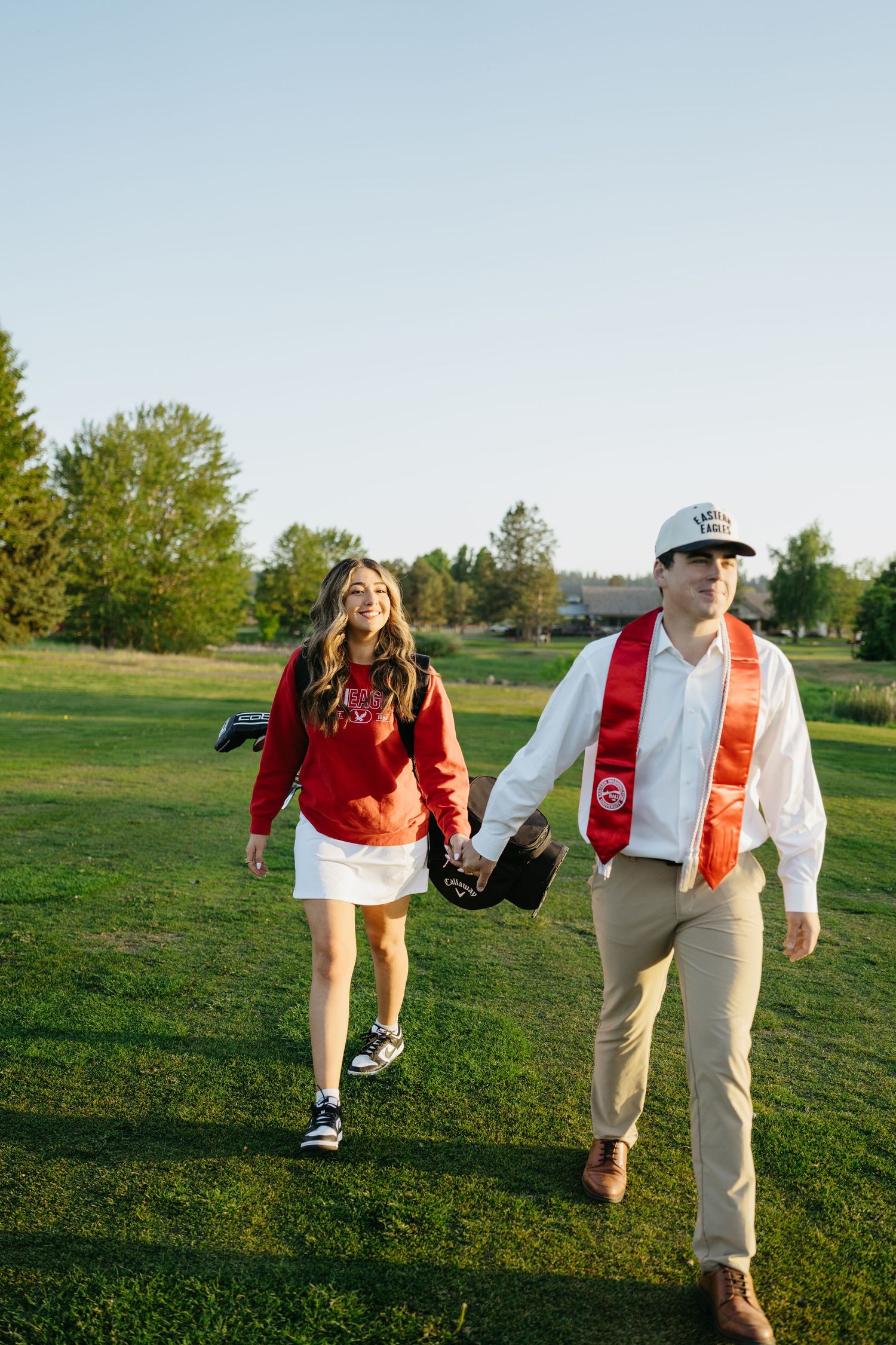A couple walks hand-in-hand on a green lawn, the man wearing a graduation stole and cap, the woman in a red sweatshirt.