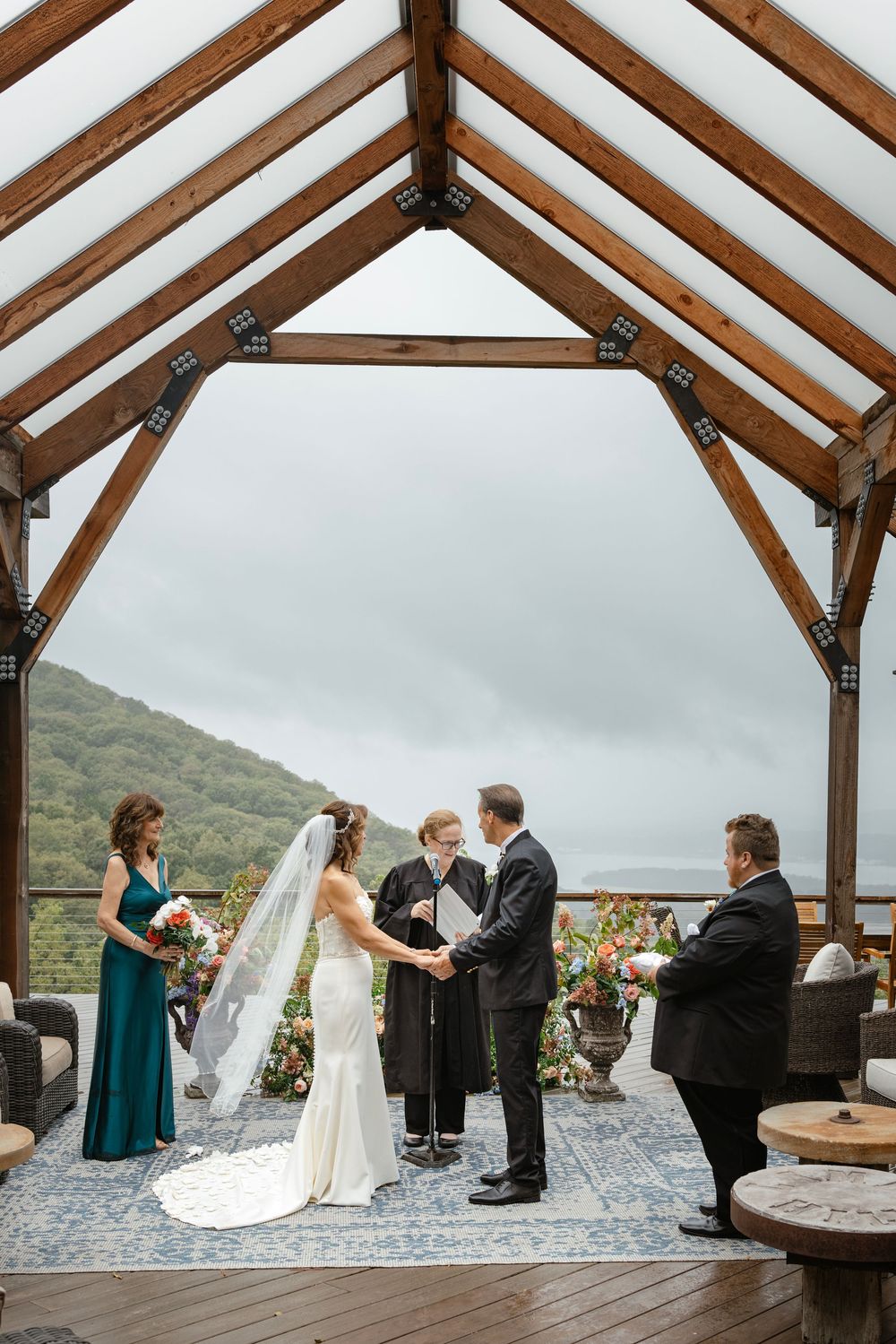 Wedding ceremony takes place under a rustic wooden pavilion overlooking mountain views on an overcast day.