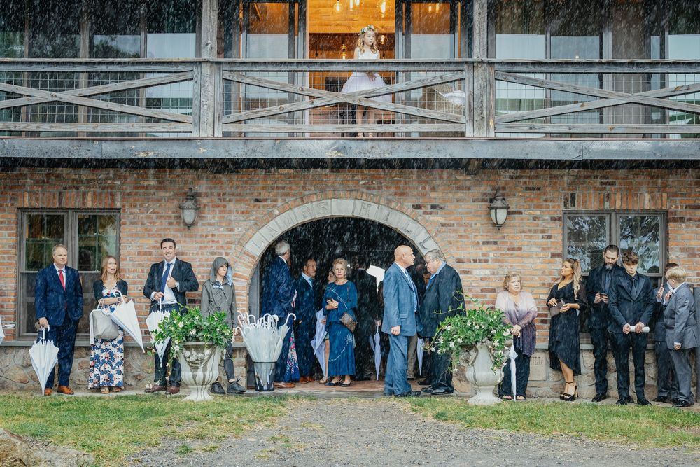 Wedding party stands in front of rustic brick wall with greenery and modern architecture in the background.