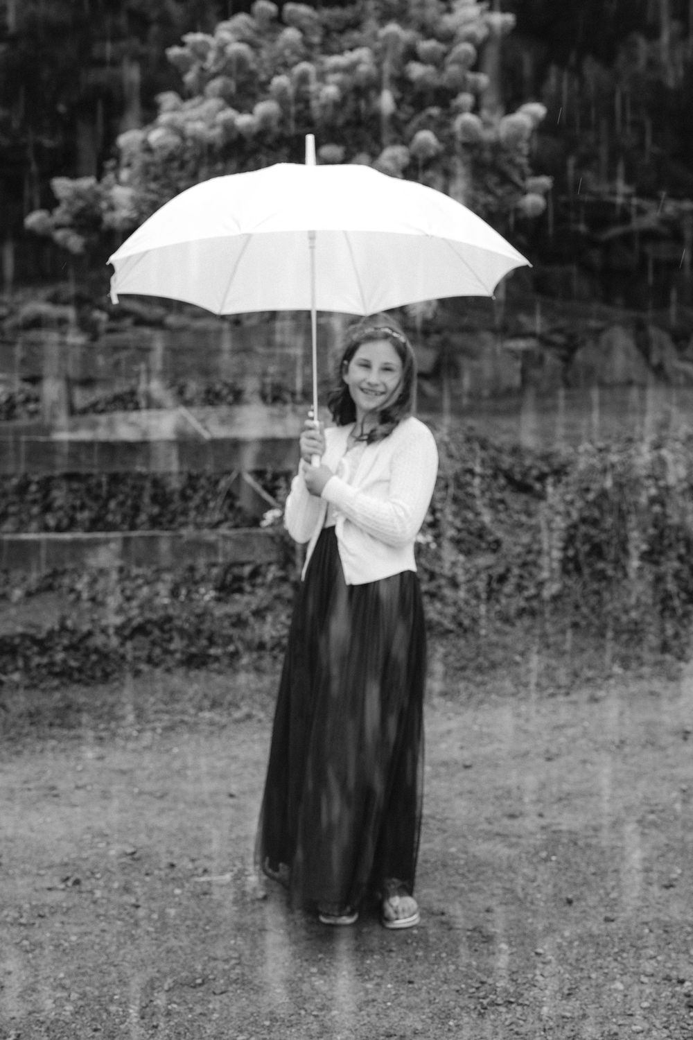 A person in dark attire stands in the rain holding a white umbrella in a garden setting.