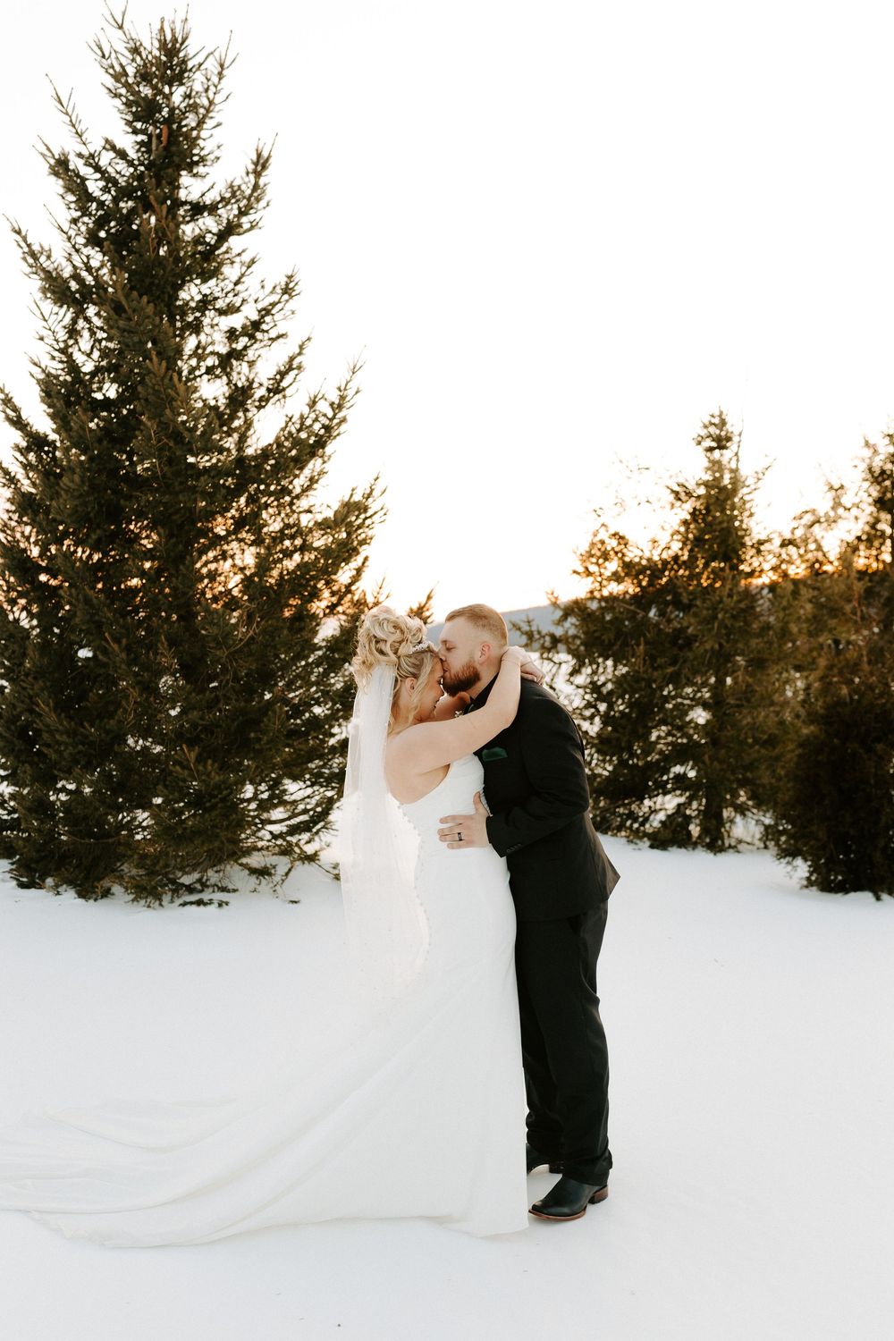 Groom lifts bride for a kiss surrounded by snow-covered pine trees at golden hour winter wedding.