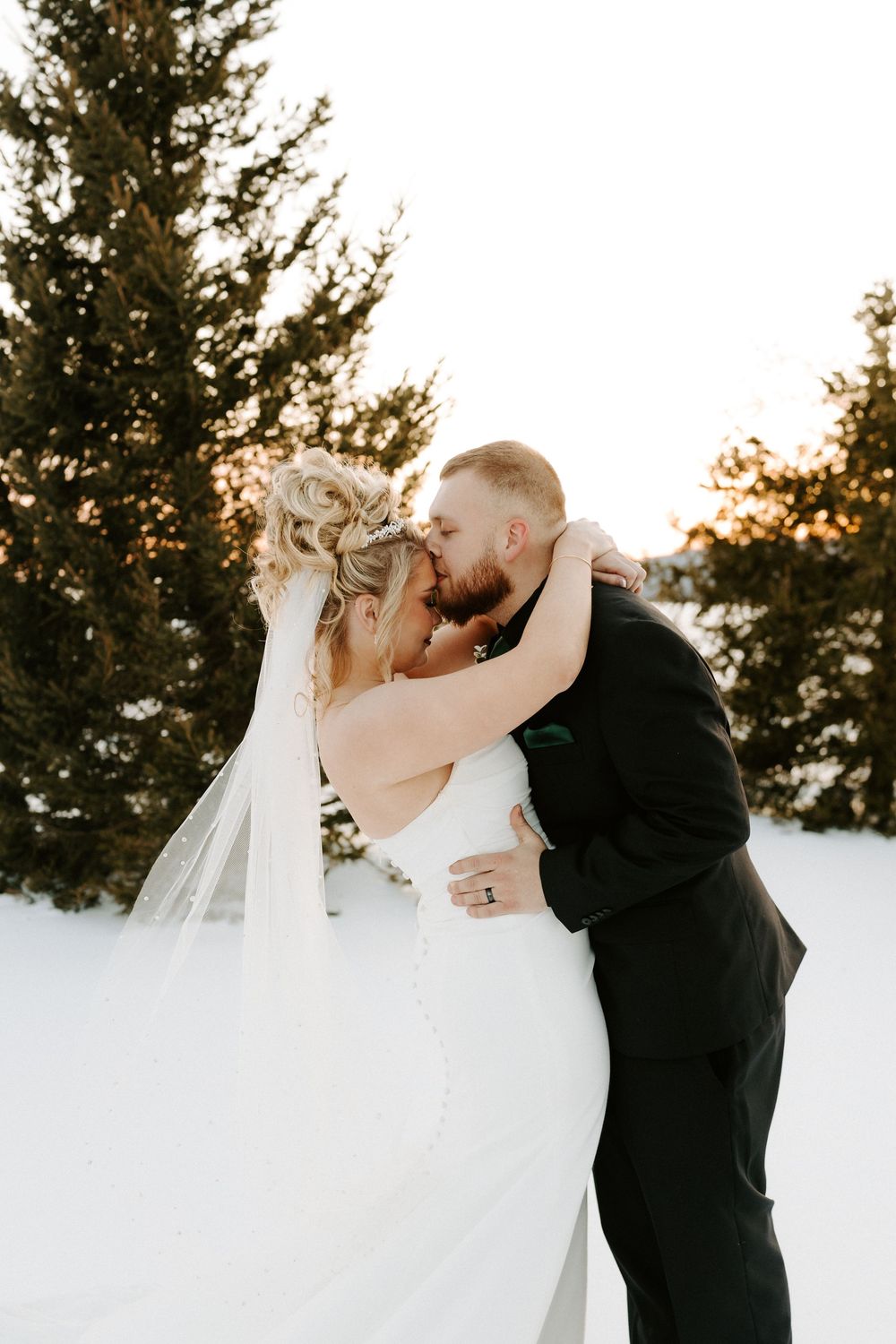 Bride and groom embrace and kiss in snowy setting with evergreen trees glowing in golden sunset light.