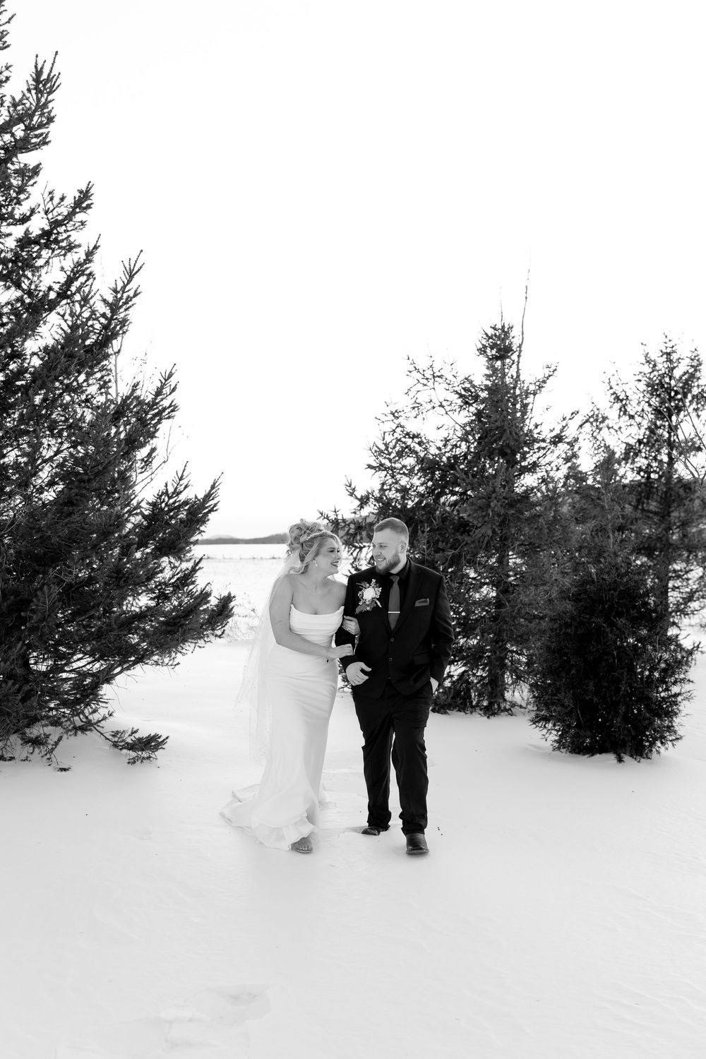 Bride and groom walk and laugh together through snowy pine forest in black and white winter wedding photo.