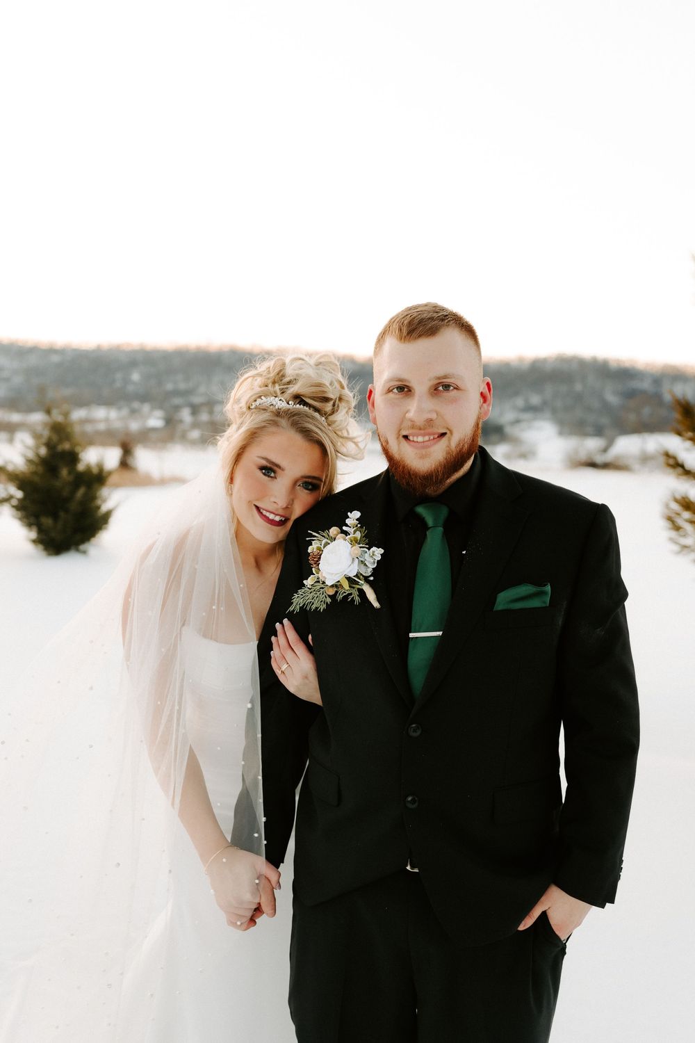 Smiling bride and groom pose together in snow, groom wearing black suit with green tie and boutonniere.