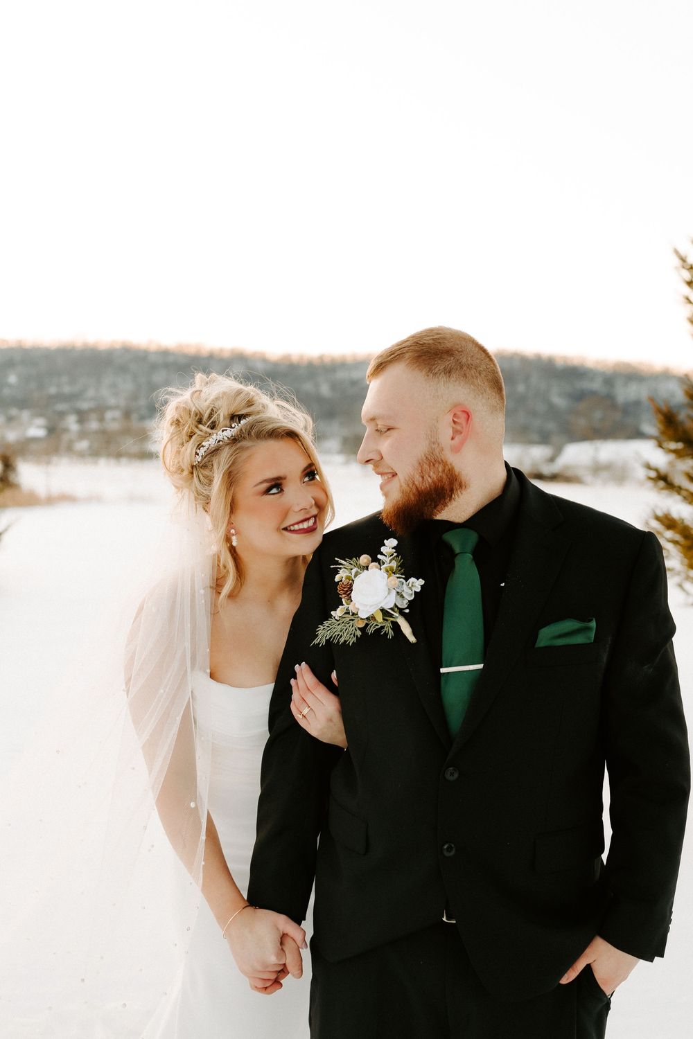 Elegant bride in white gown and veil walks with groom in black suit through snowy winter scenery.