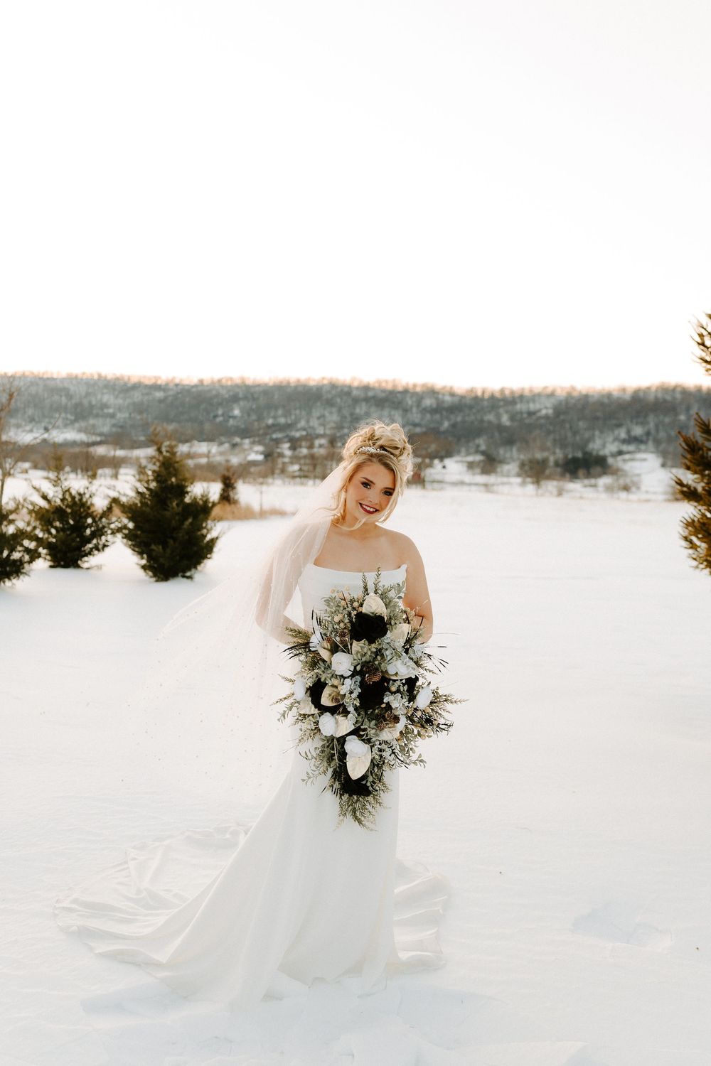 Bride in strapless white gown holds a dramatic cascading bouquet of winter blooms in a snowy landscape.