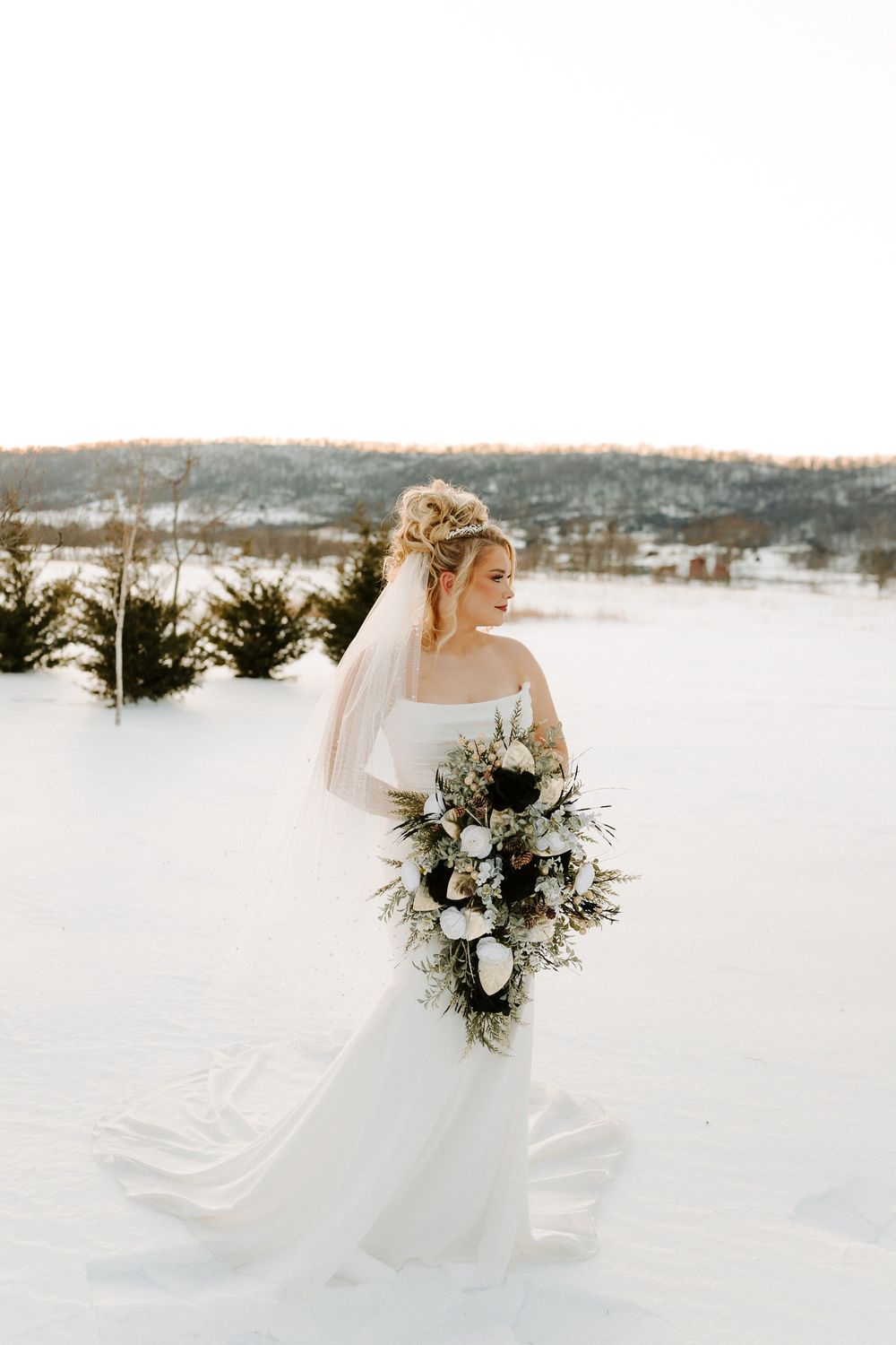 Elegant bride in flowing white gown and veil carries a lush cascading bouquet in a winter snow setting.