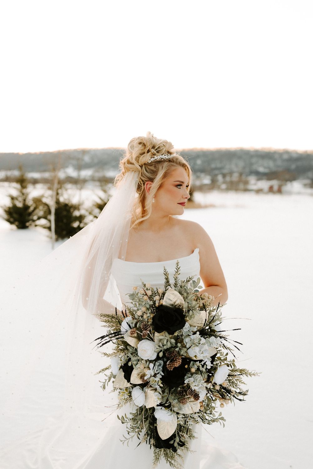 Bride in strapless gown holds a stunning cascading winter bouquet with dark and white blooms in the snow.