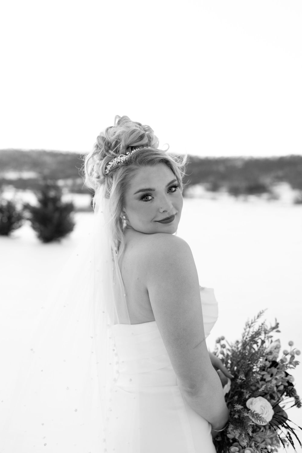 Black and white portrait of a bride glancing over her shoulder holding a winter bouquet in snowy scenery.
