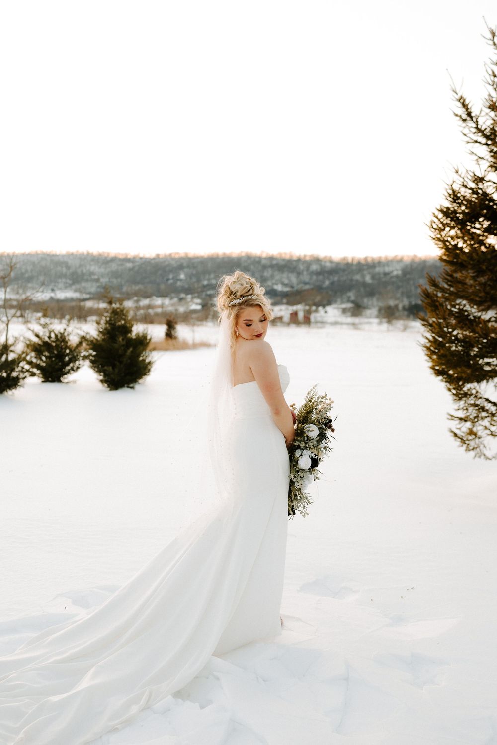 Bride in white strapless gown with veil holding bouquet in snowy winter landscape.
