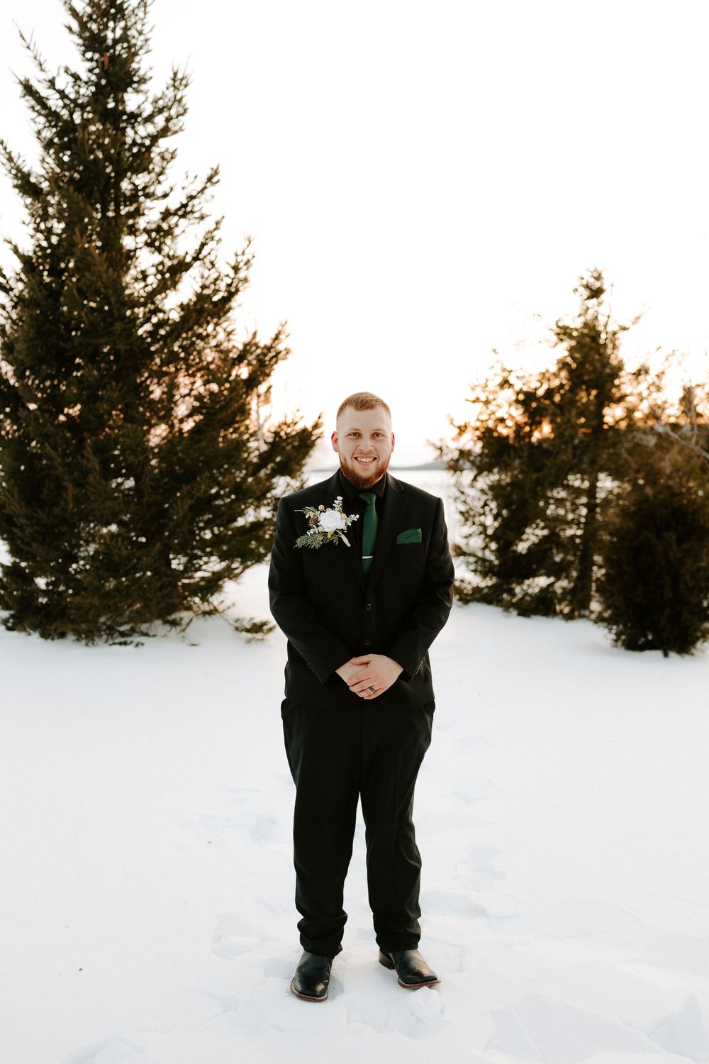 Groom in black suit with green tie and boutonniere standing in snowy winter setting.