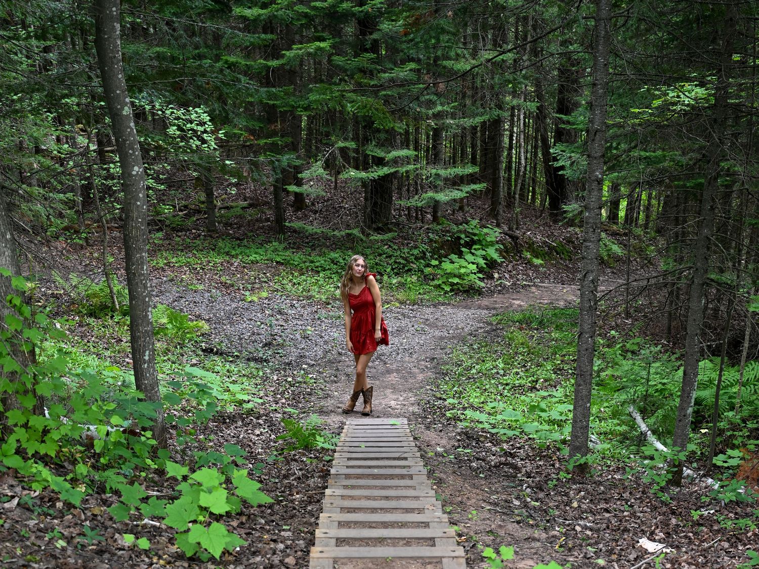 Woman in red dress standing on wooden boardwalk steps in a lush green forest trail.