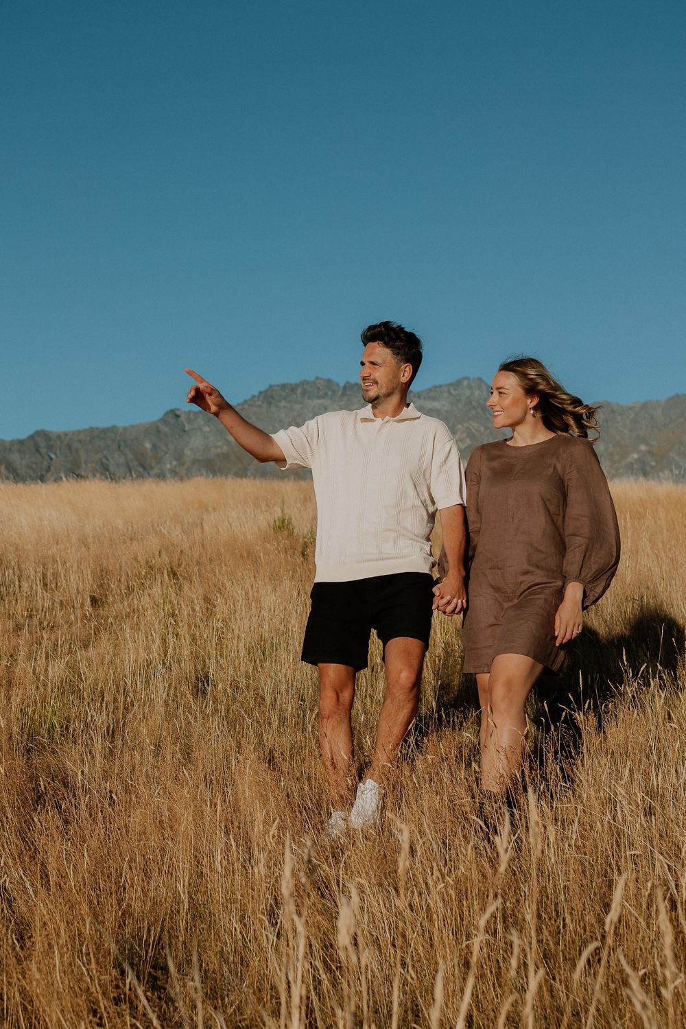 Two people walking through a golden grass field against a mountain backdrop in warm sunlight.