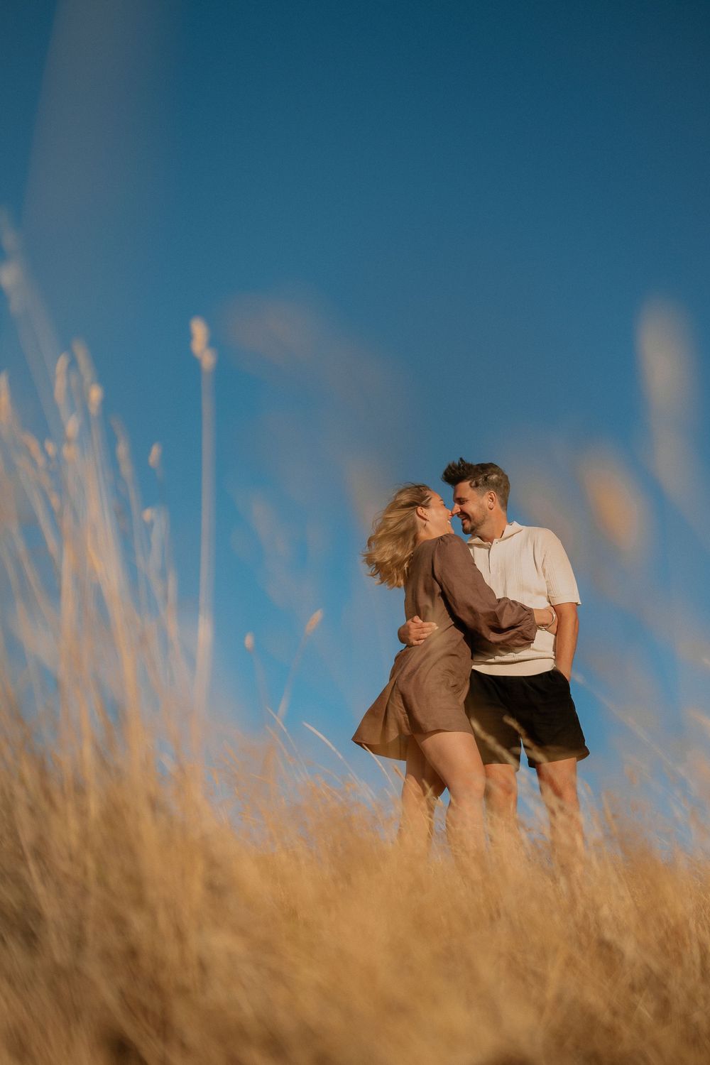 A couple shares an intimate moment together while standing in golden wheat field against blue sky at sunset.