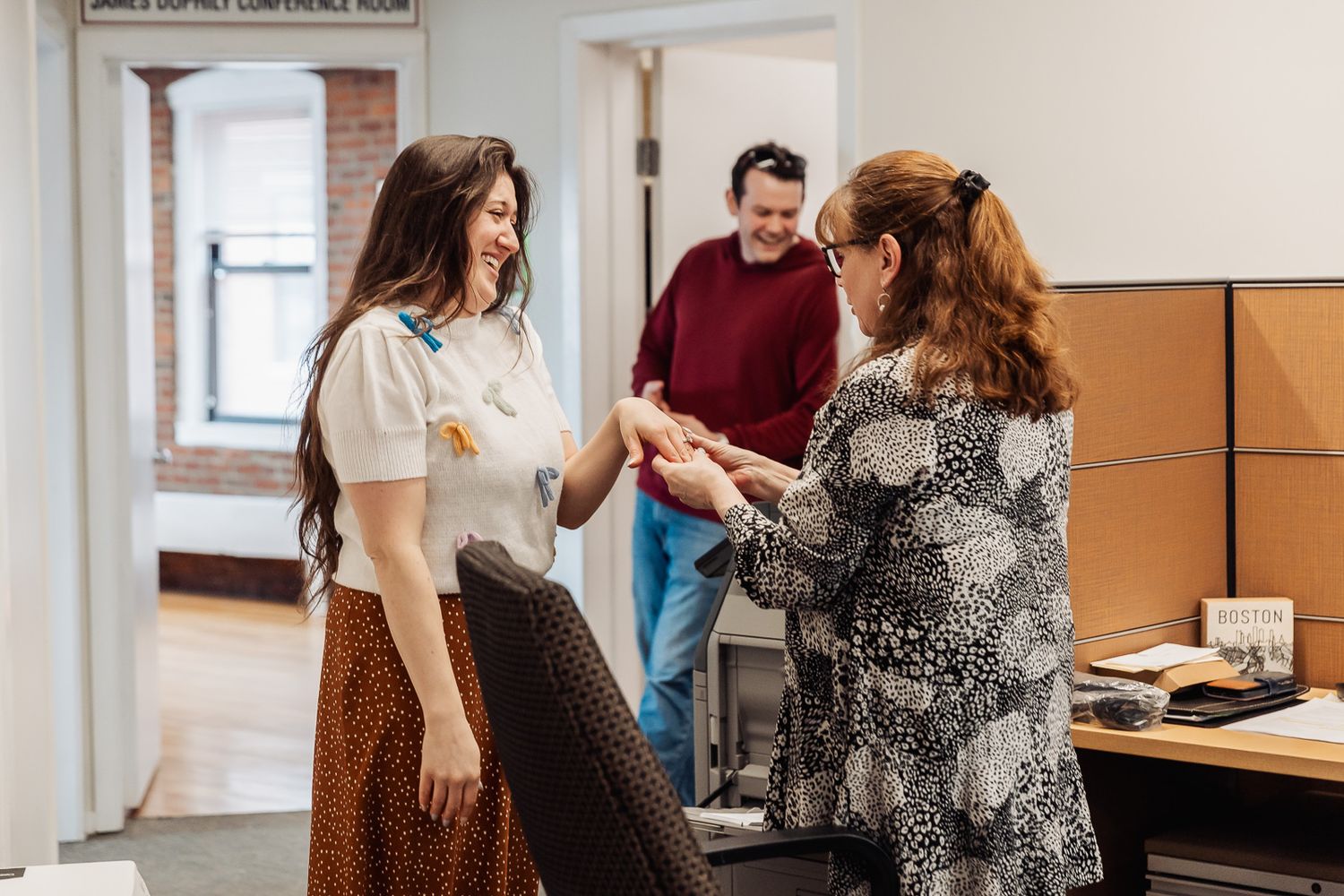 Two women shaking hands and smiling in a modern office, with a man standing in the background.