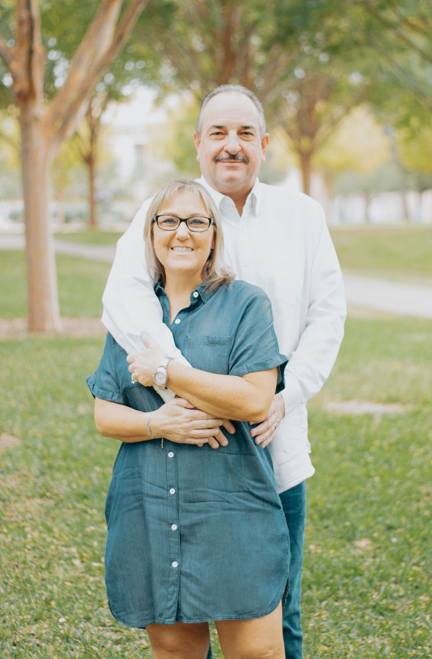 A couple embraces while standing outdoors in a park with trees in the background.