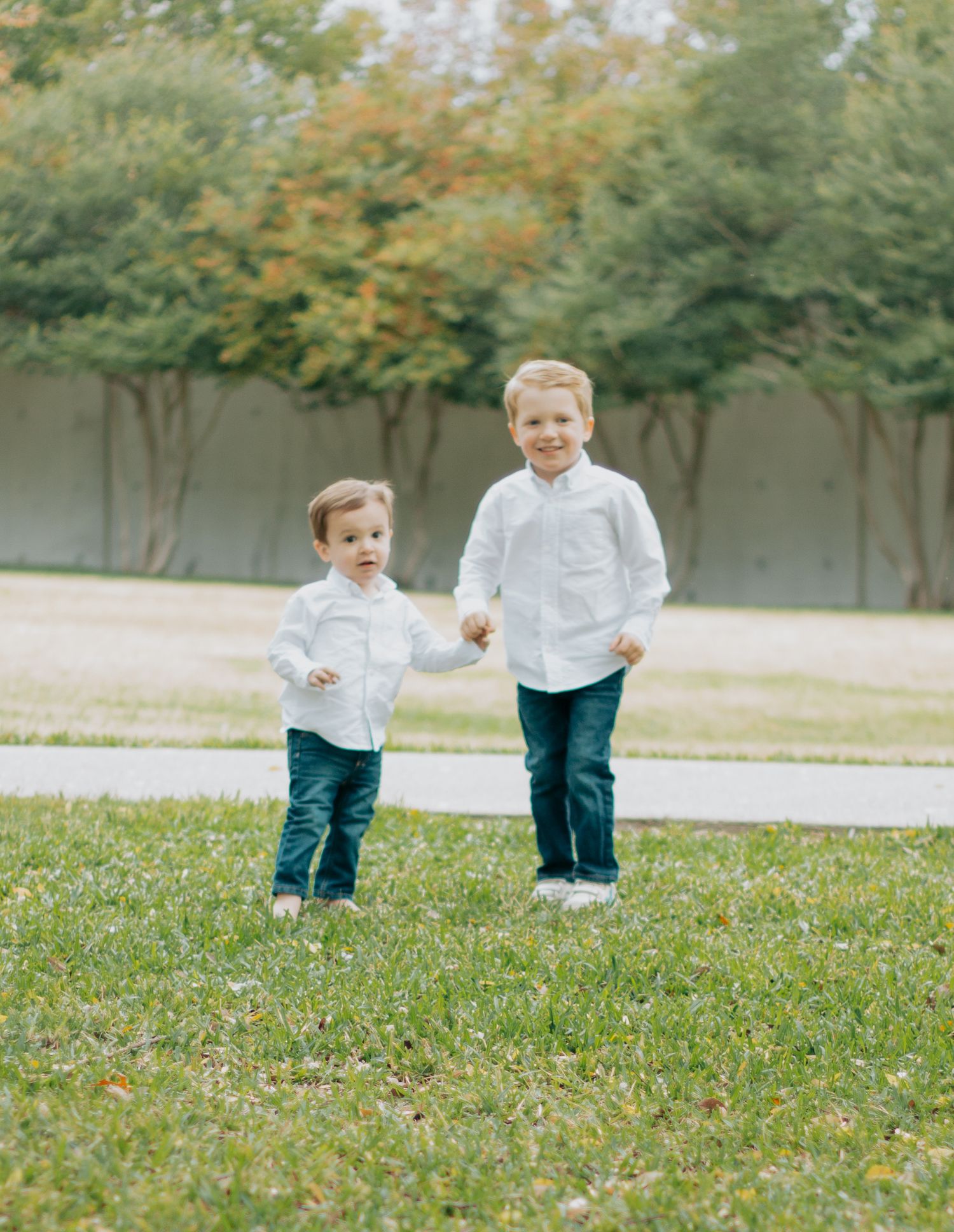 Two children in white shirts and blue pants stand together on grass with autumn trees in the background.