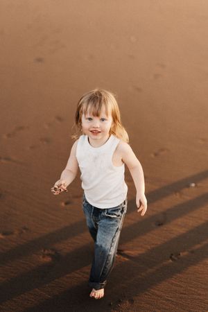 Small child in white tank top and jeans running joyfully on sand.
