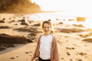 Child standing on beach at golden sunset wearing brown cardigan over white shirt.
