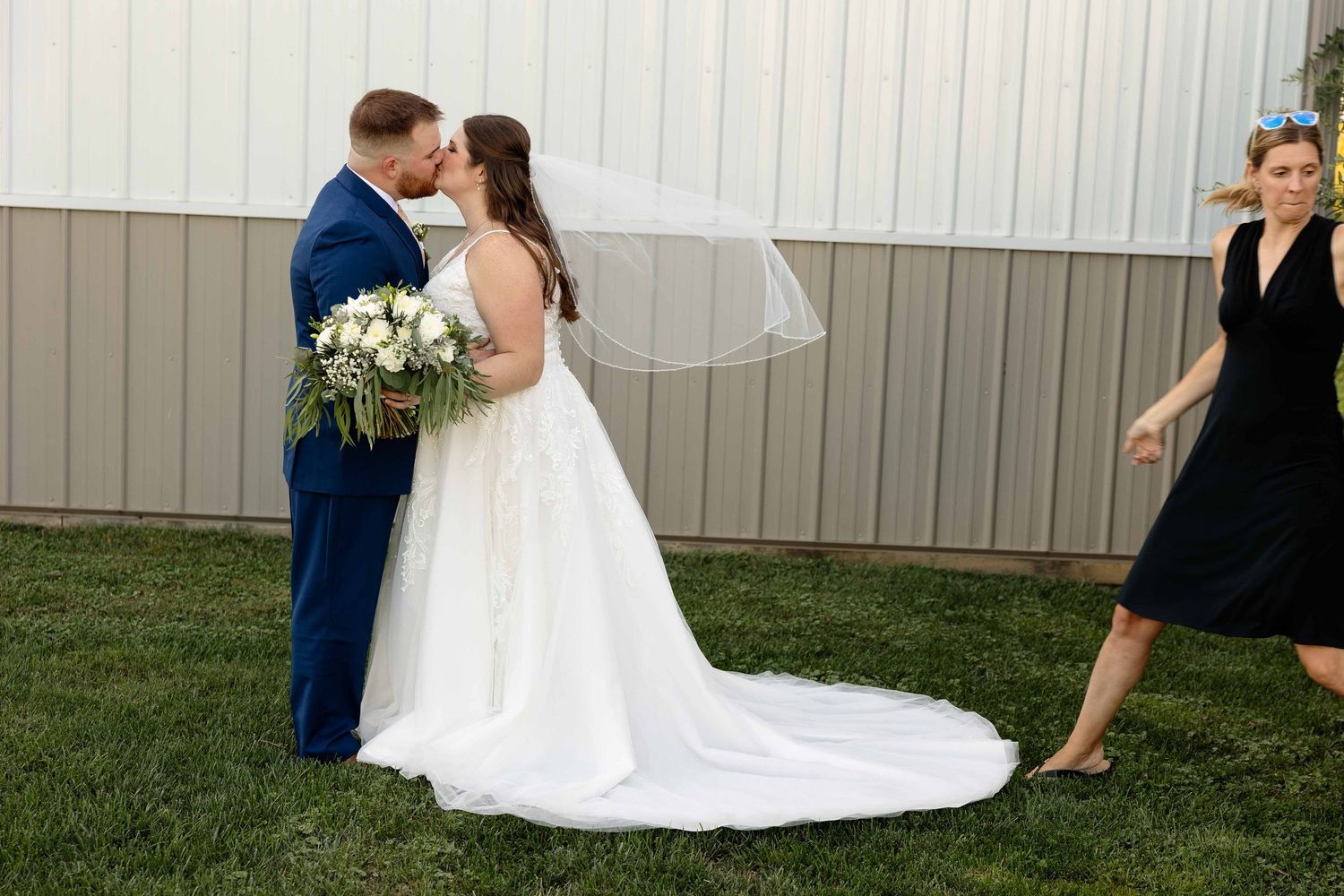 A wedding couple shares a romantic kiss during their outdoor ceremony next to a white barn wall.