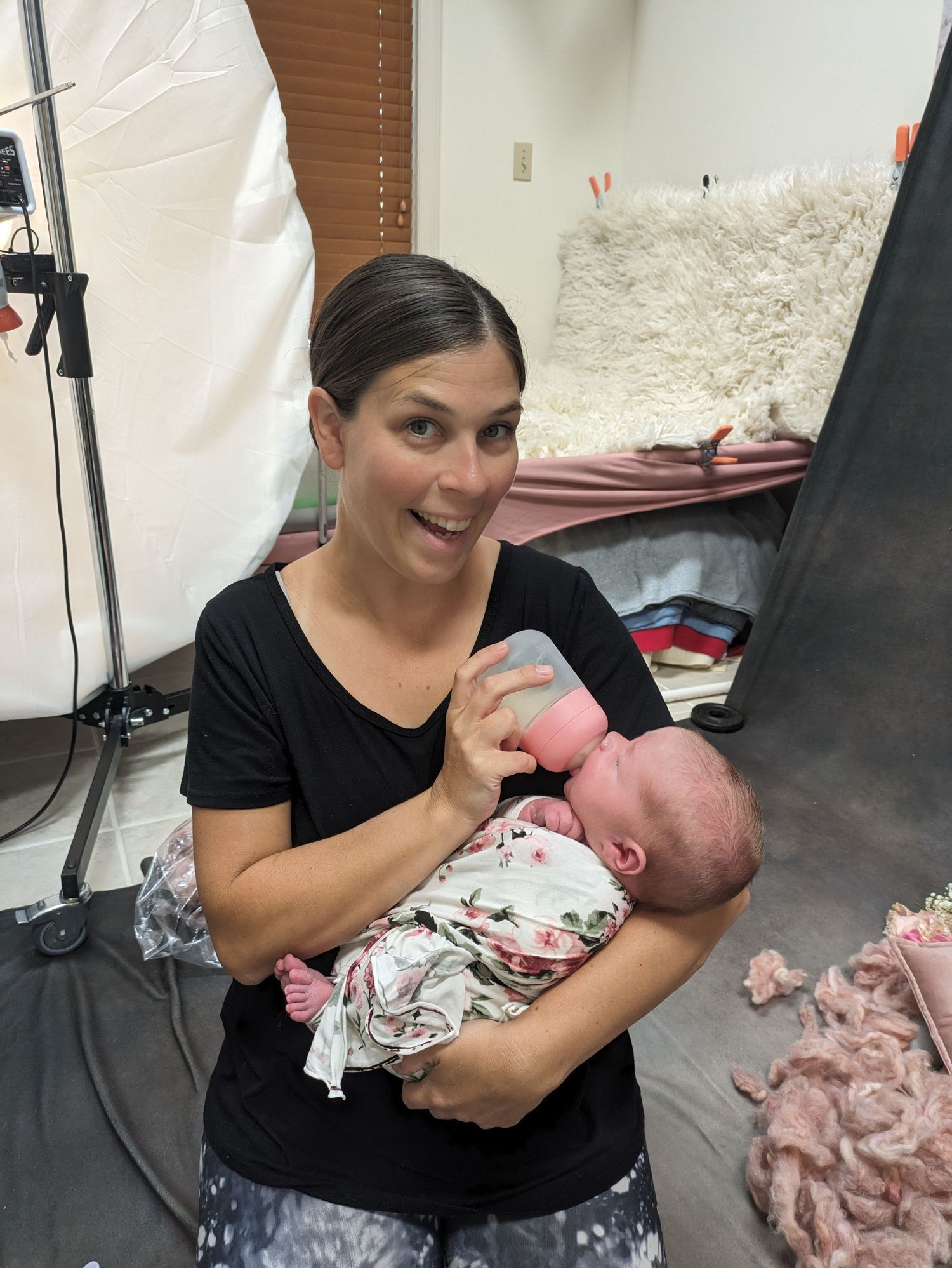 A person in a black shirt holds and feeds a swaddled newborn baby in what appears to be a photography studio setting.