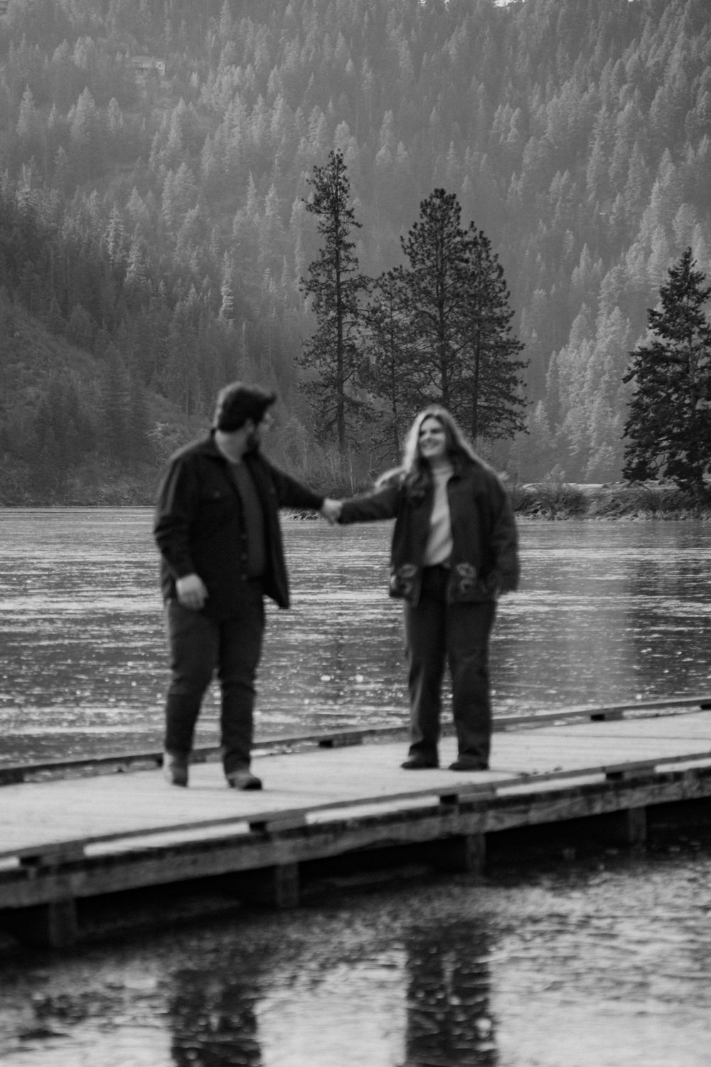 Two people standing on a wooden dock by a lake surrounded by pine trees in a snowy, forested landscape.