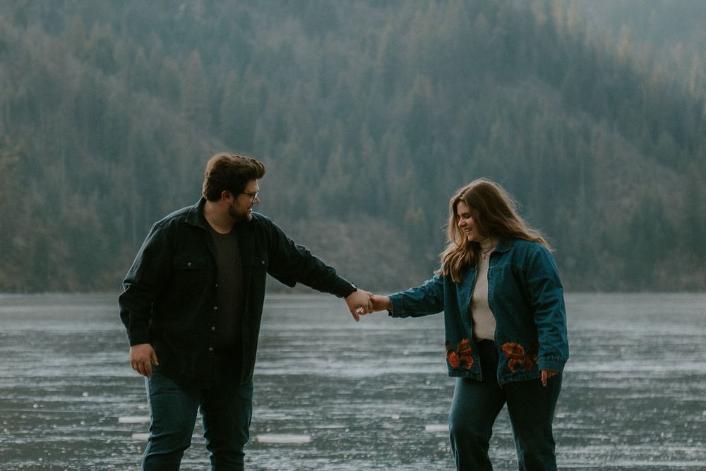 Two people standing in a body of water, with a forested landscape in the background.