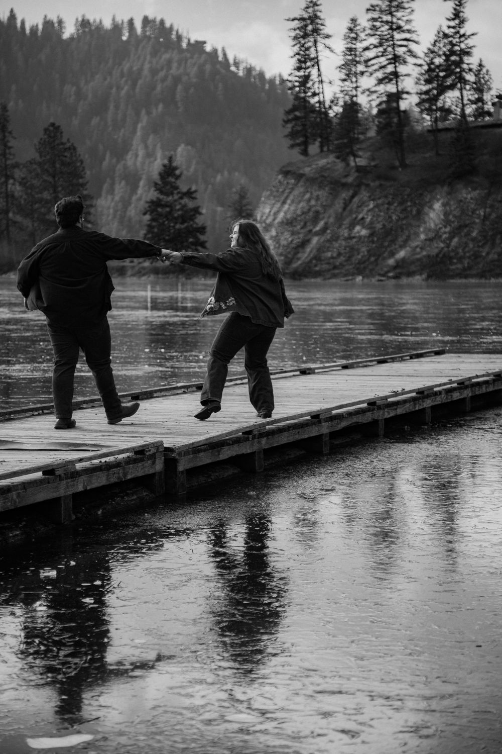 Two people standing on a wooden dock overlooking a scenic river surrounded by forested mountains.