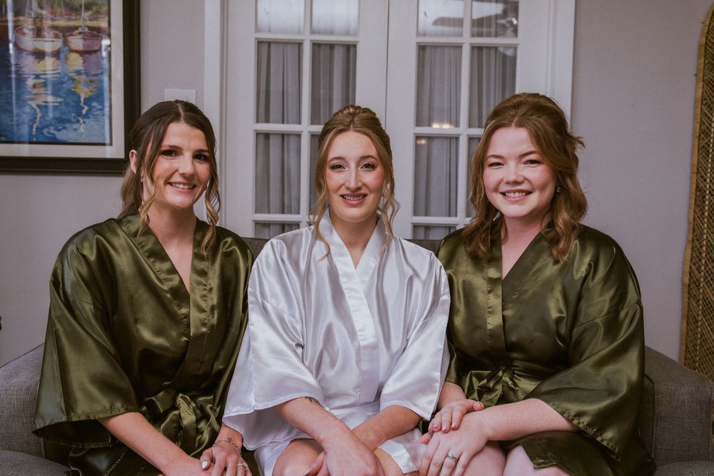 Three women in satin robes smile together, two in olive green and one in white, likely a bride with bridesmaids.