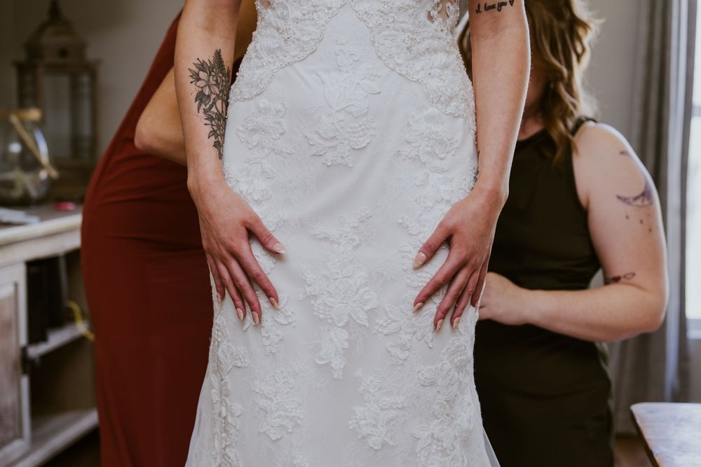 Two bridesmaids in burgundy and olive dresses adjusting a lace wedding gown, both with floral tattoos.