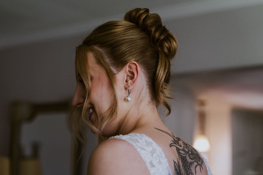 Bride with elegant updo hairstyle, pearl drop earring, and floral shoulder tattoo wearing a white lace wedding dress.