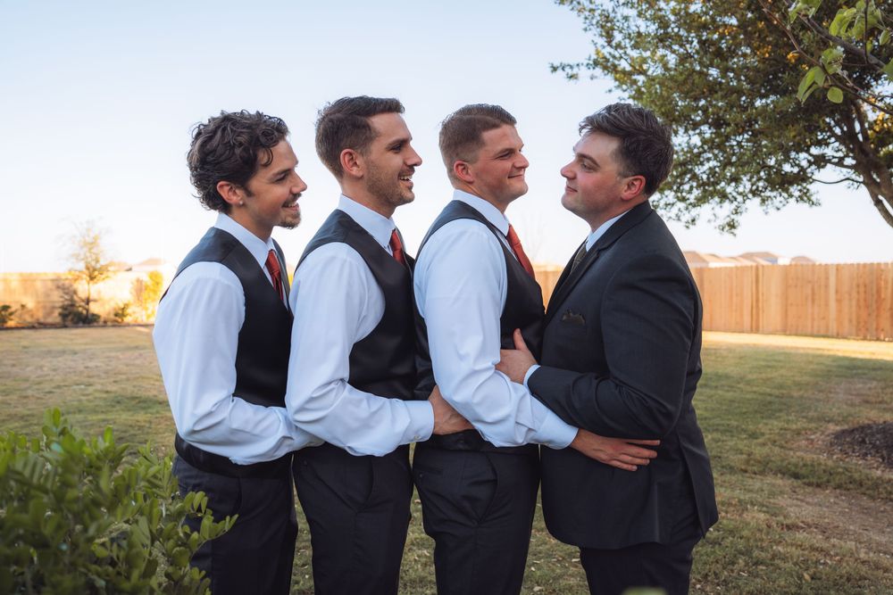Four groomsmen in dark suits and red ties posing and smiling together in a sunny outdoor setting.