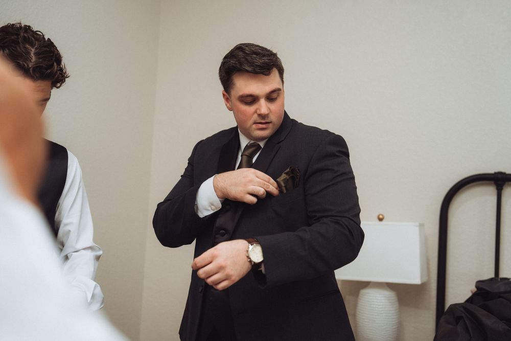 Groom adjusting cufflinks on dark suit before wedding ceremony indoors.