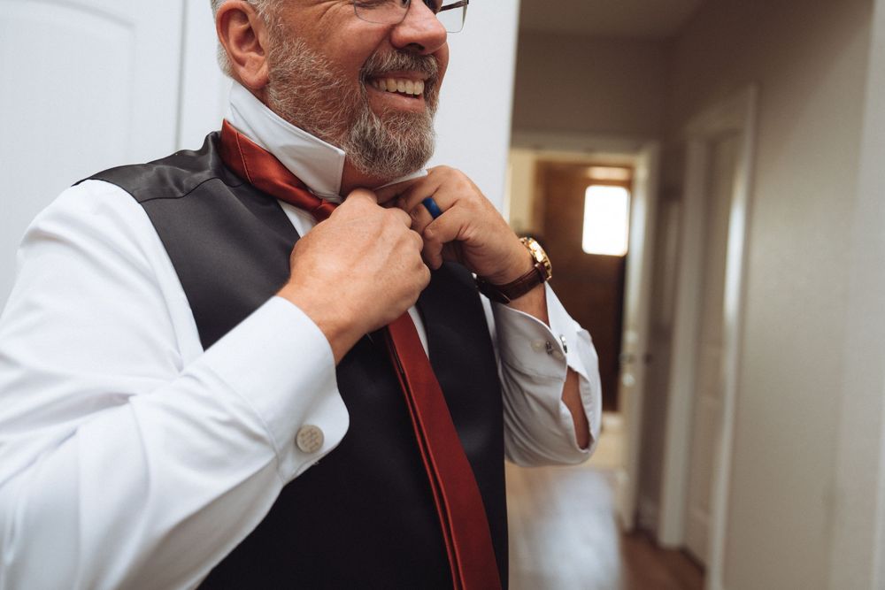Smiling middle-aged man in vest and white shirt adjusting red tie, getting dressed for formal occasion.