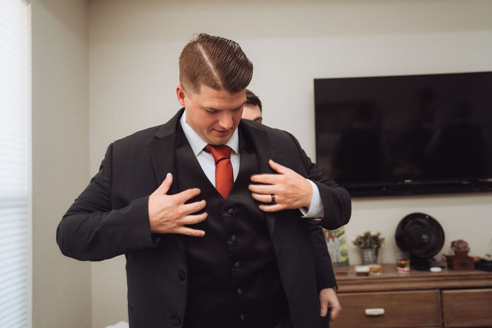 Groom in black suit and red tie adjusting his jacket before wedding ceremony.