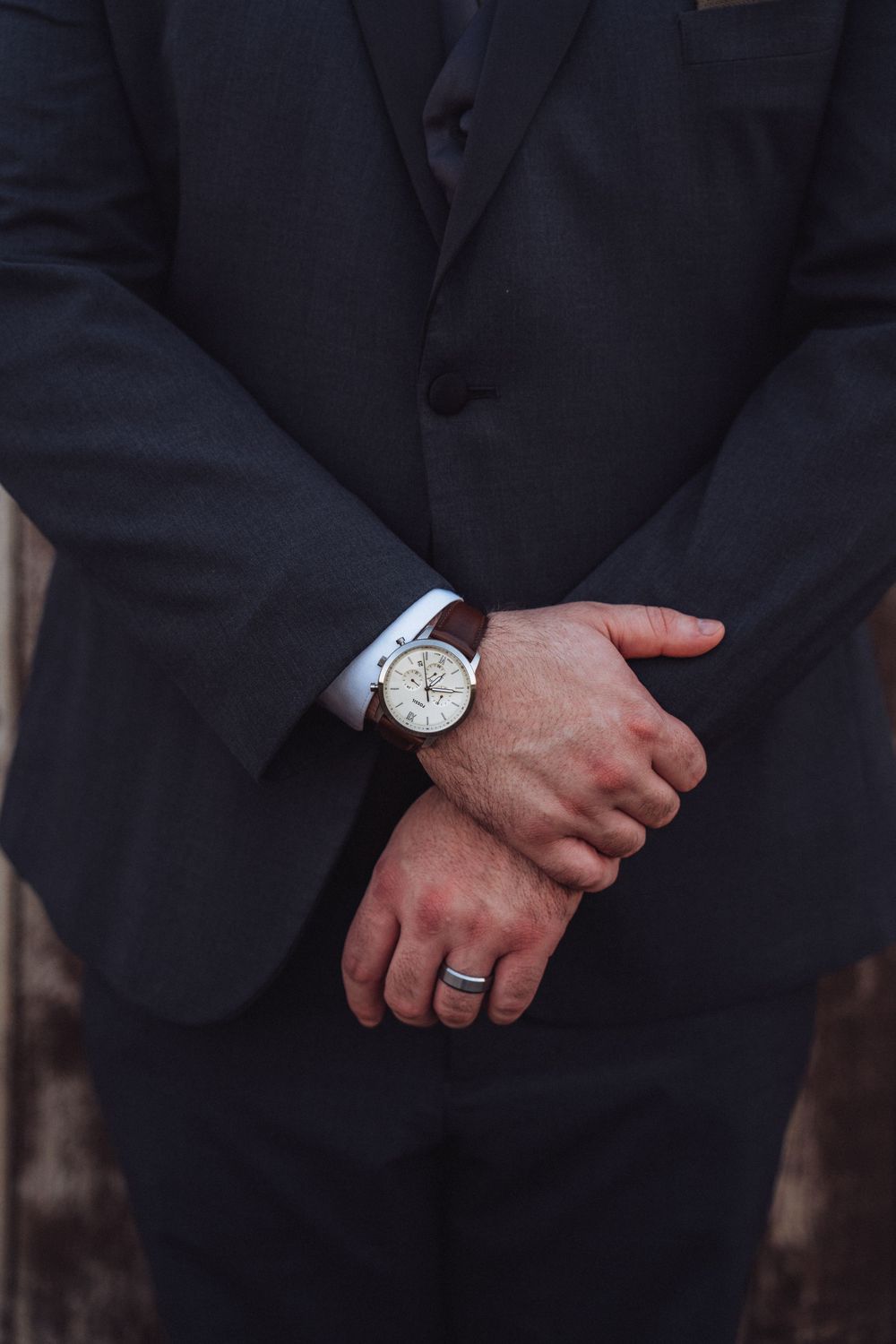 Groom in dark navy suit with clasped hands wearing a silver watch and wedding band close-up.
