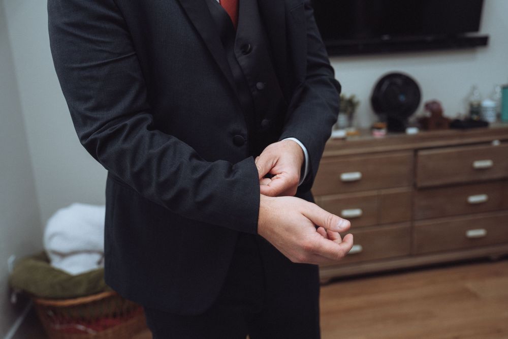 Groom in black three-piece suit with red tie fastening cufflinks before wedding.