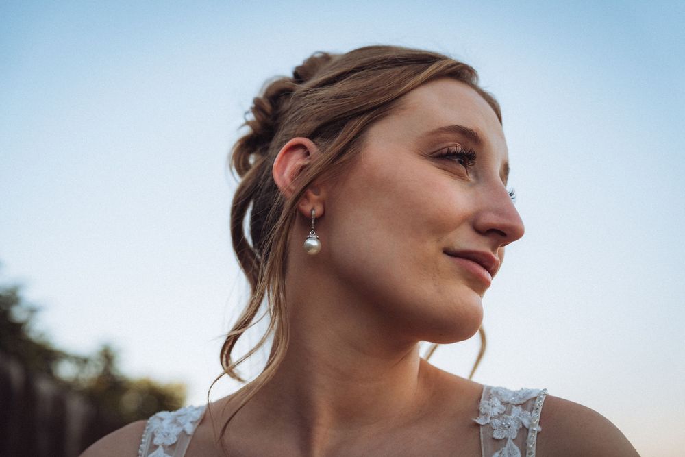Beautiful bride with updo hairstyle wearing pearl drop earrings smiling softly against a blue sky.